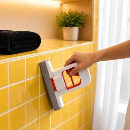 A hand using a white and orange mini mop  to clean yellow wall tiles in a bathroom setting.